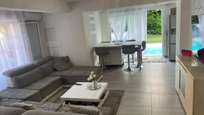 Een moderne woonkamer met een modulaire bank, een salontafel en een eethoek met een tafel en stoelen. Er is ook een keuken met een koelkast en een televisie.