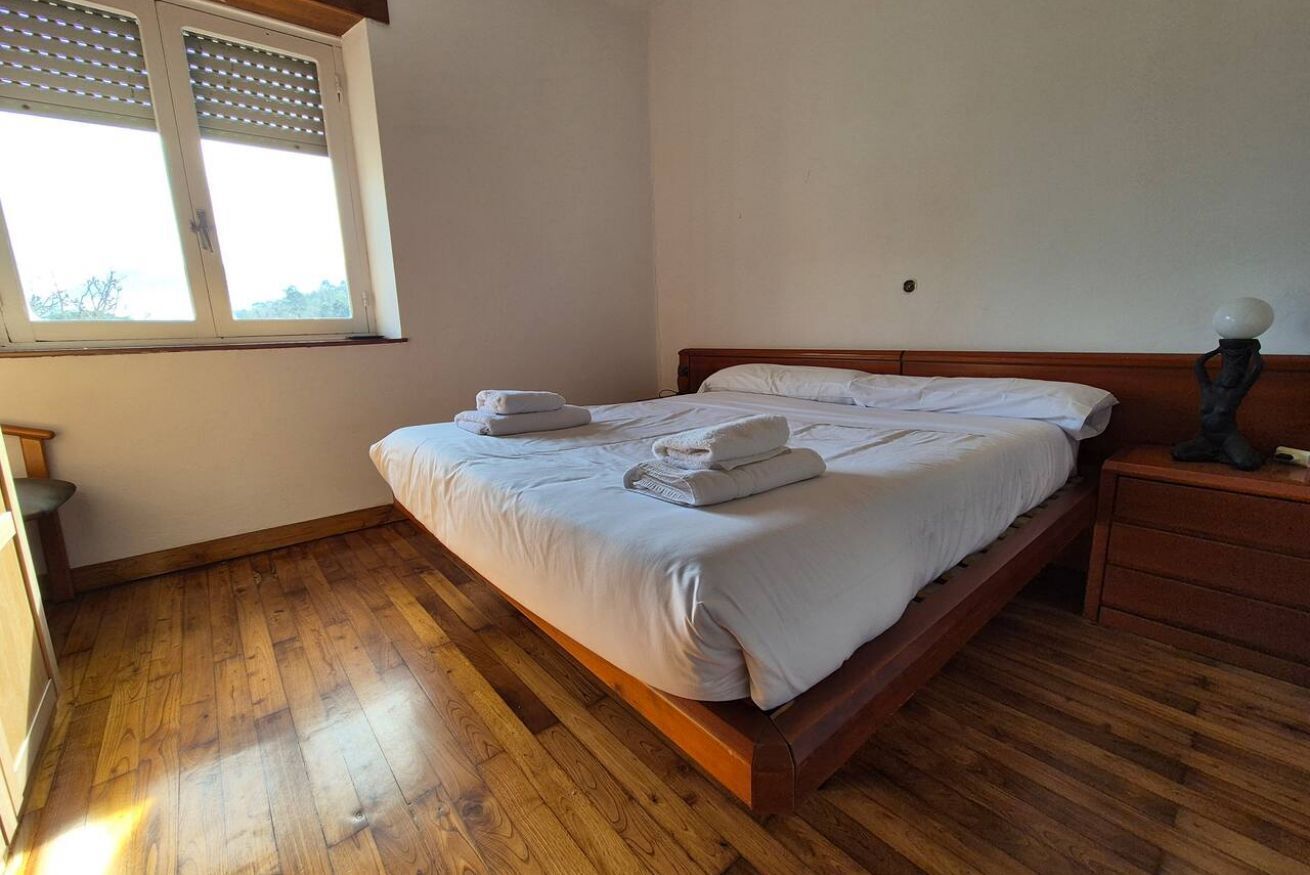 A bedroom with a wooden bed, white linens, a nightstand, a lamp, and a window with blinds.
