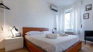 Una cama bien hecha con sábanas blancas y toallas enrolladas, una mesita de noche con lámpara, una silla negra y una ventana con cortinas.