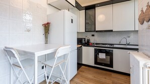 Una cocina compacta con armarios blancos, un refrigerador blanco, una estufa y un lavamanos. Hay un pequeño comedor con una mesa y sillas blancas.
