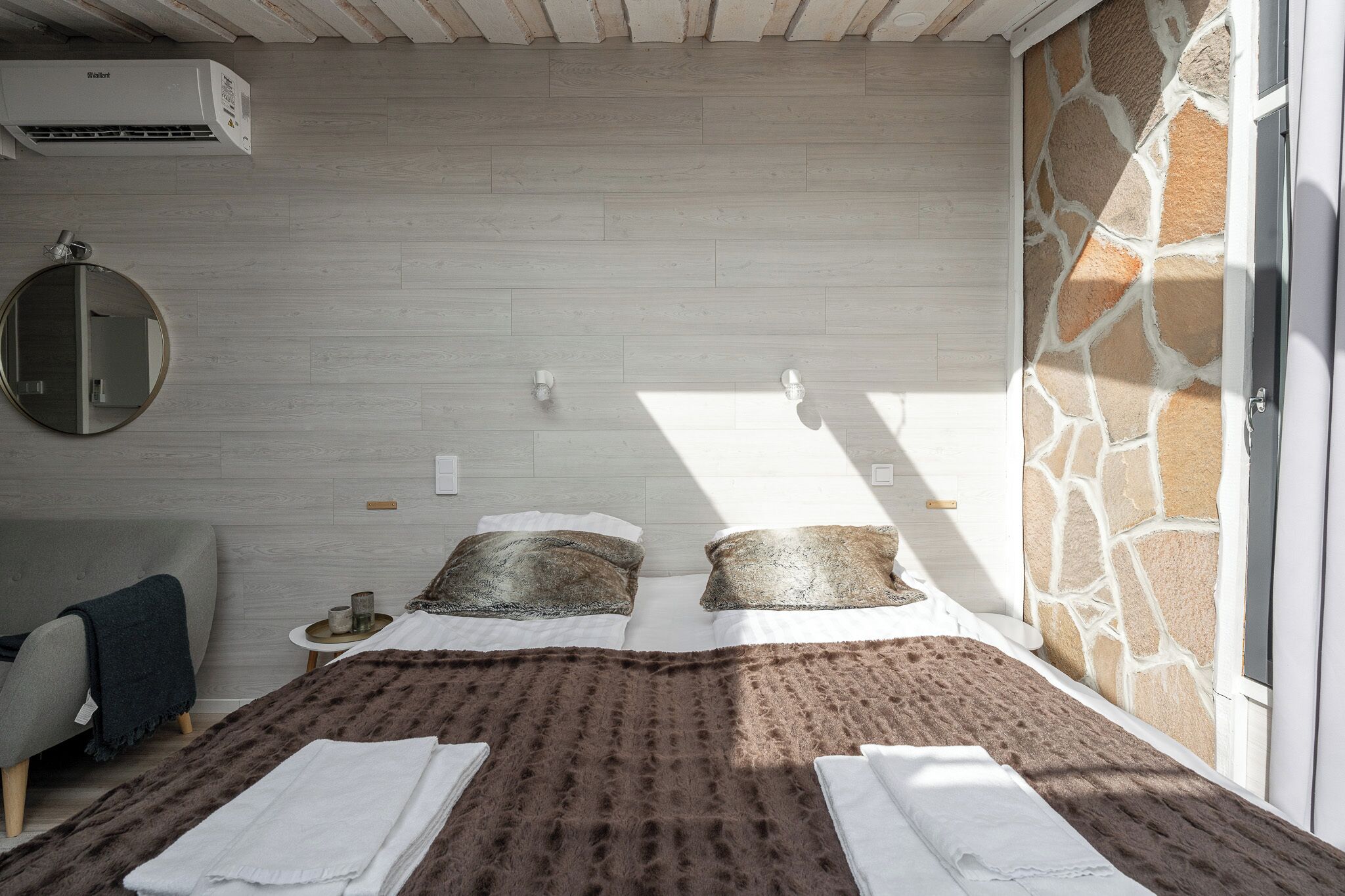 A bedroom with a bed, a round mirror, a small table with a candle, and a stone wall.