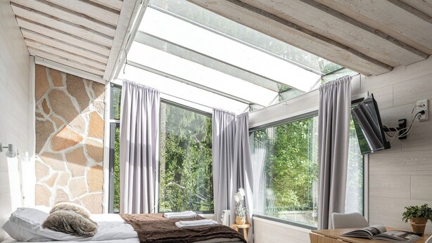 A modern room with a bed, sofa, and a small table, large windows with curtains, and a stone wall. - Koskisviitti 1 (Laukaa)