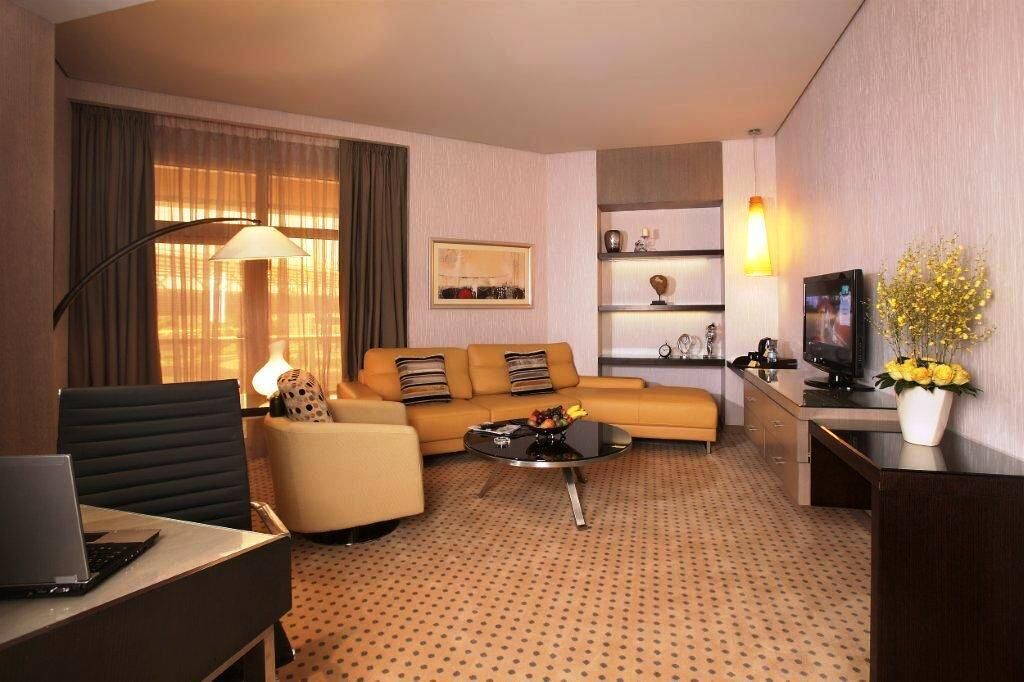 A hotel room with a sofa, armchair, coffee table, TV, and a desk with a laptop.