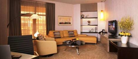 A hotel room with a sofa, armchair, coffee table, TV, and a desk with a laptop.