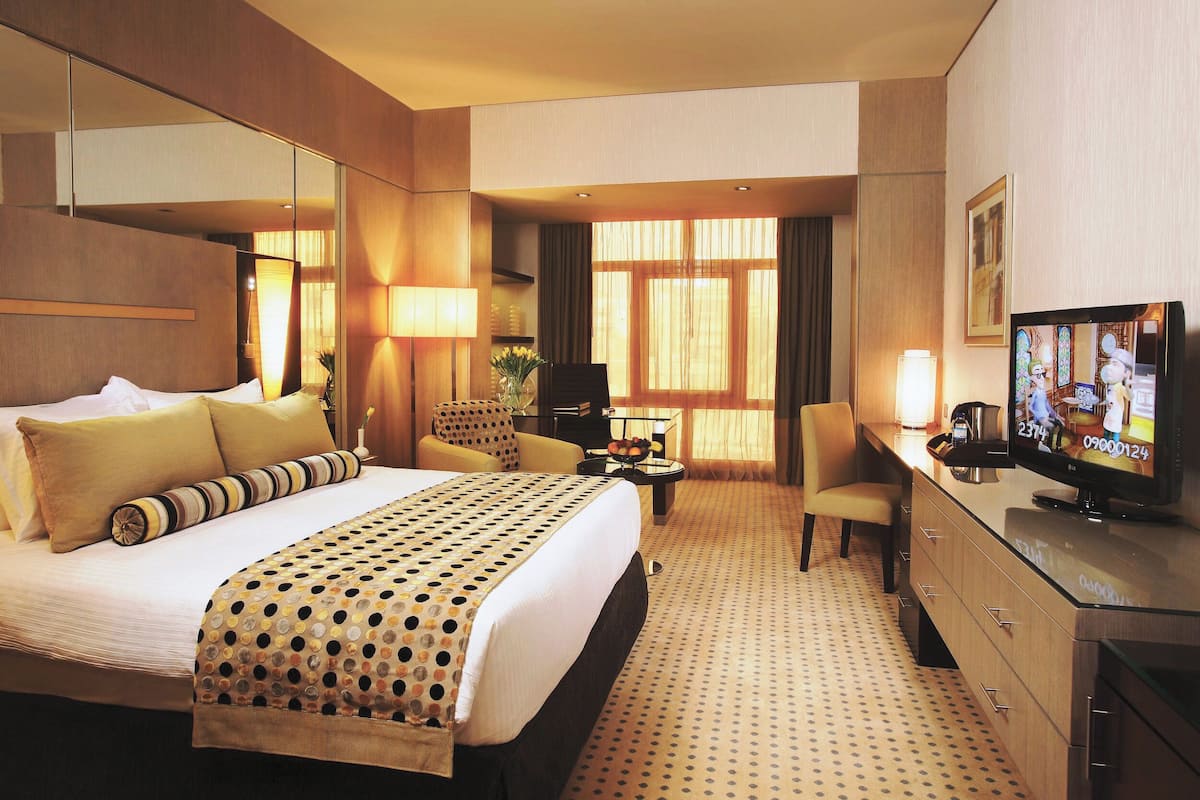 A hotel room with a large bed, a desk with a television, a chair, and a desk lamp.