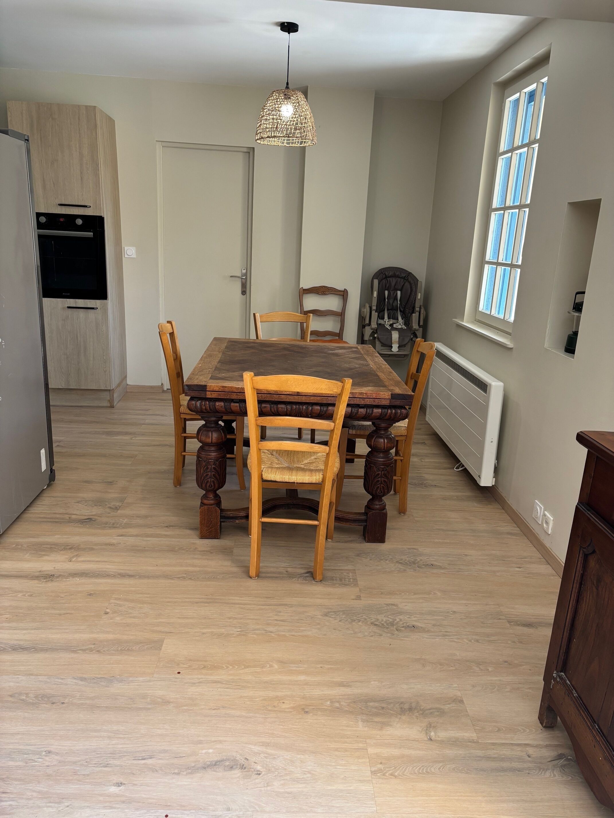 Un comedor con una mesa y sillas de madera, una lámpara colgante, una ventana con persianas y un radiador.