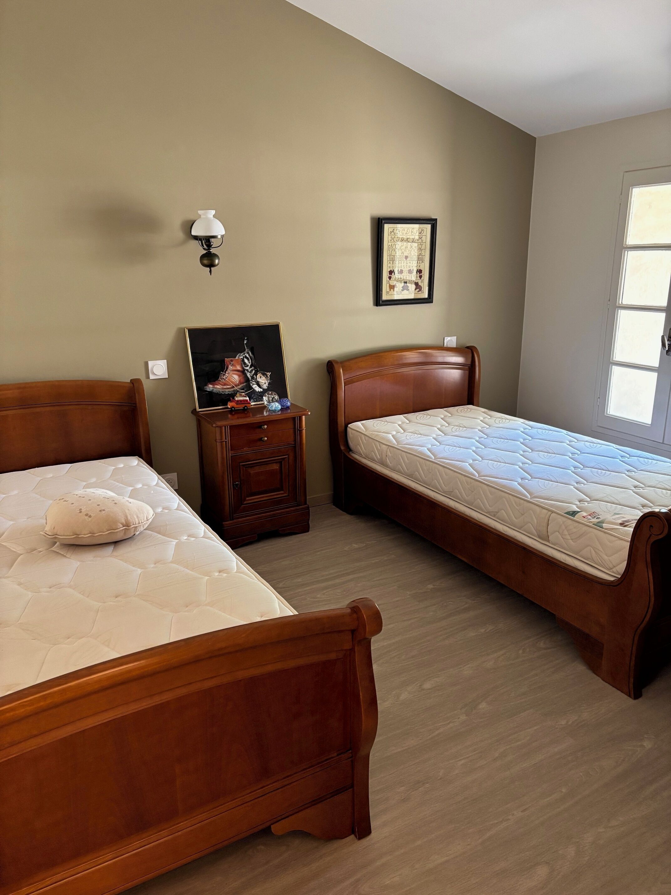 A bedroom with two wooden beds, a nightstand with a lamp, a framed picture, and a window.