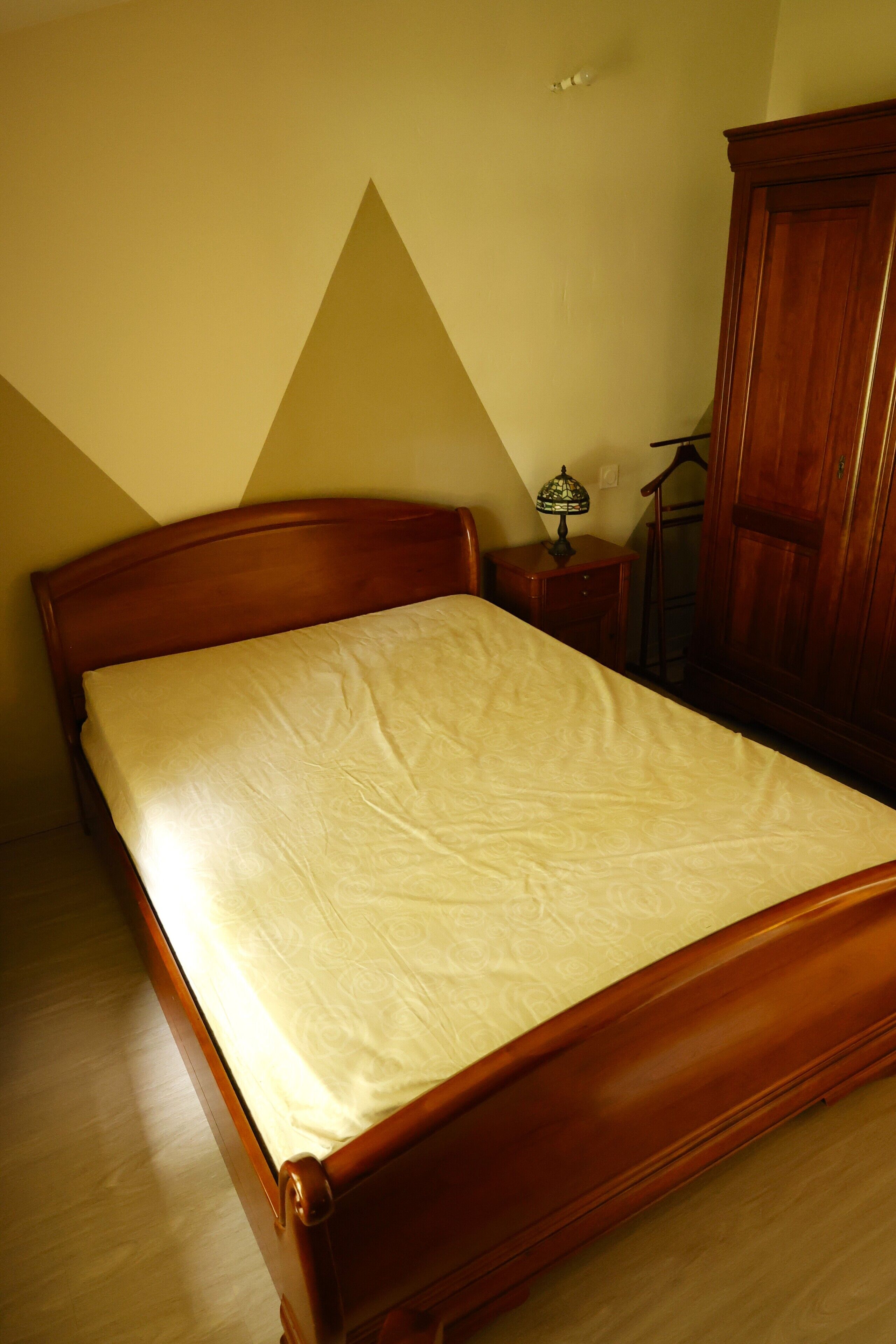 A neatly made bed in a room with a wooden headboard, a small bedside table with a lamp, and a wardrobe.