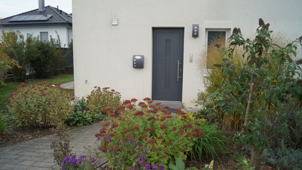 Ett modernt hus med en grå dörr, en liten trädgård med olika växter och en stenlagd gång som leder till entrén.