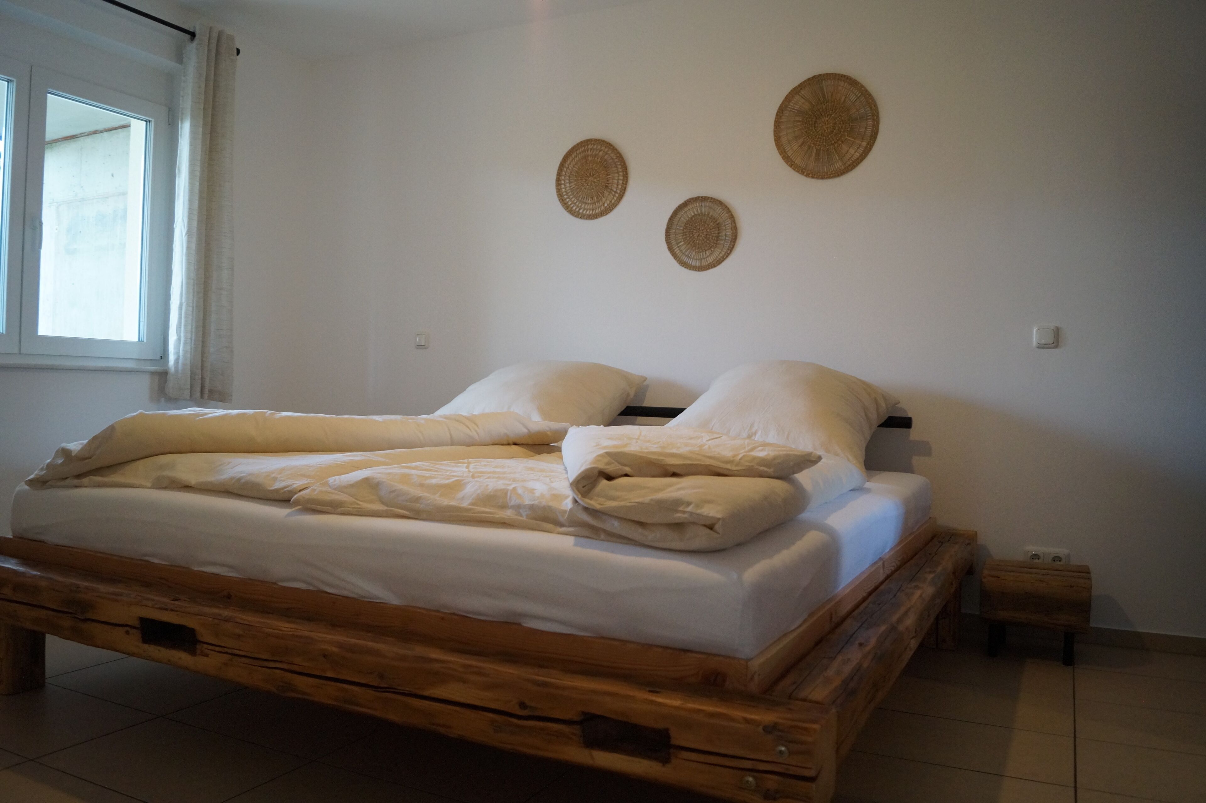 Un letto di legno con biancheria da letto bianca in una stanza dalle pareti bianche e con una finestra.