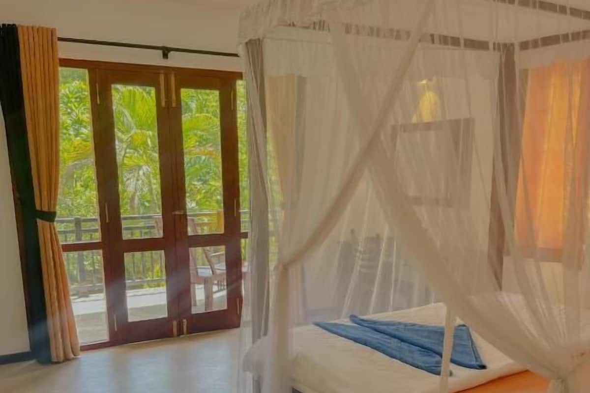 Un dormitori amb un llit sota una bugada, un ventilador de sostre i vistes a la vegetació a través de les portes de vidre.