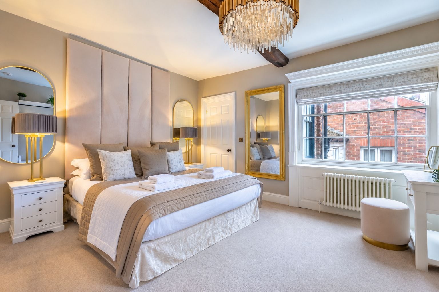 Une chambre à coucher avec un grand lit, des lampes de chevet, un miroir, un radiateur et une fenêtre donnant sur un immeuble en briques.
