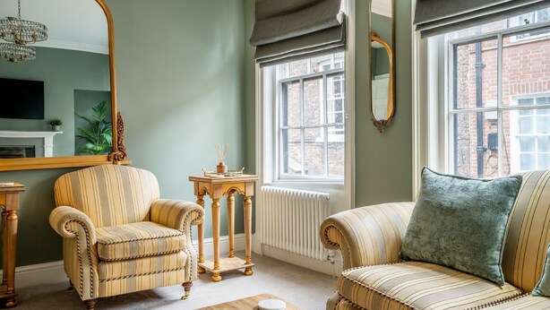 A living room with a sofa, armchair, and a large mirror. There is a window with a view of a brick building outside. - Friargate House -  a premium stay that sleeps 9 guests  in 4 bedrooms (York)