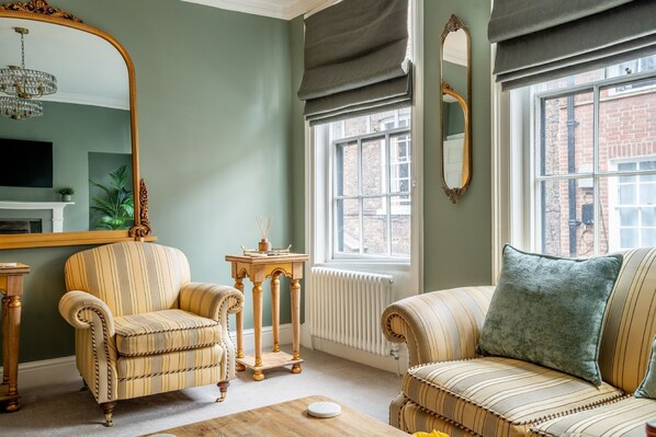 Een woonkamer met een bank, fauteuil en een grote spiegel. Er is een raam met uitzicht op een bakstenen gebouw.