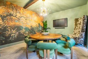 Un espace repas comprenant une table ronde en bois, des chaises à revêtement vert, une grande fresque murale, un lustre moderne et un téléviseur à écran plat fixé au mur.