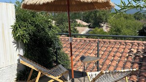A wooden deck with two lounge chairs and a thatched umbrella, overlooking a tiled roof and greenery.