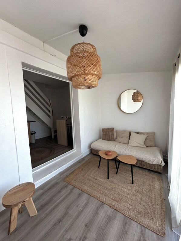 Een gezellige woonkamer met een bank, twee bijzettafels, een geweven vloerlamp en een ronde spiegel aan de muur.