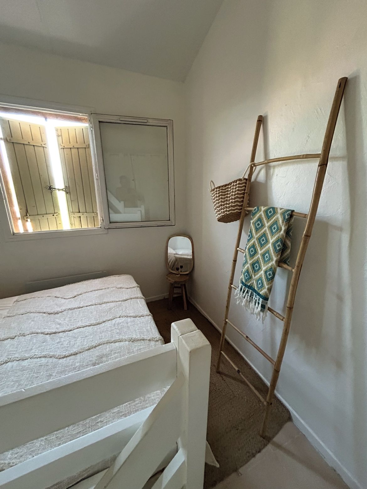 Quarto pequeno e bem arrumado, com uma cama, janela com portadas, espelho, escada de madeira e um banquinho.