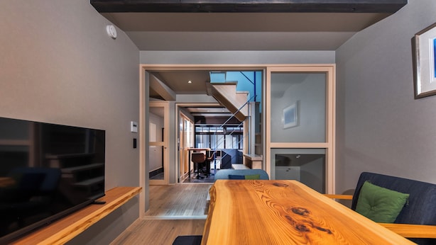 Un interior moderno con una mesa de comedor de madera, un televisor de pantalla plana sobre un soporte de madera, y una escalera que conduce a un nivel superior.