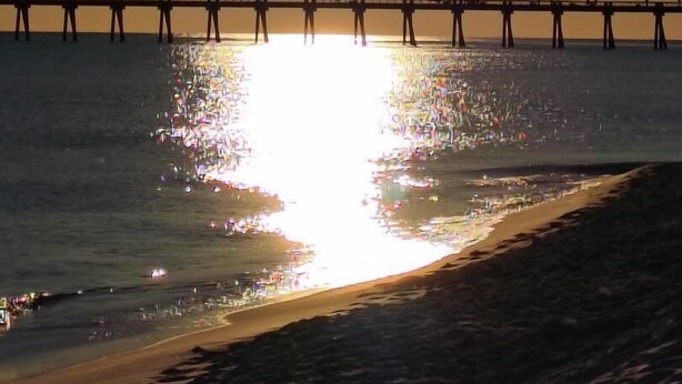 Un muelle se adentra en aguas tranquilas, con un sol brillante reflejado en la superficie.
