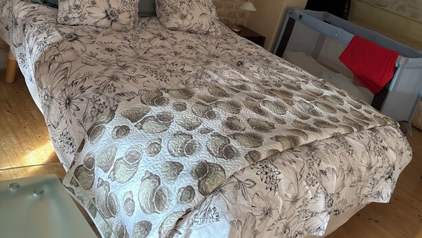 Camera da letto con parete in pietra, travi in legno, un letto con biancheria a fiori e una valigia sul pavimento.