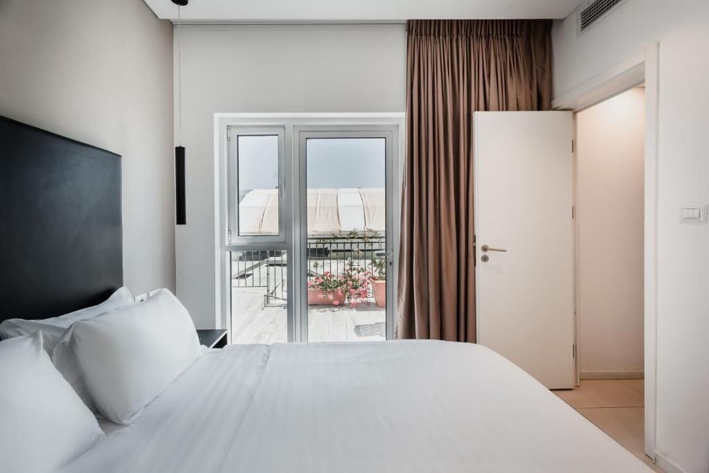 A hotel room with a bed, a door, and a view of a balcony with plants.