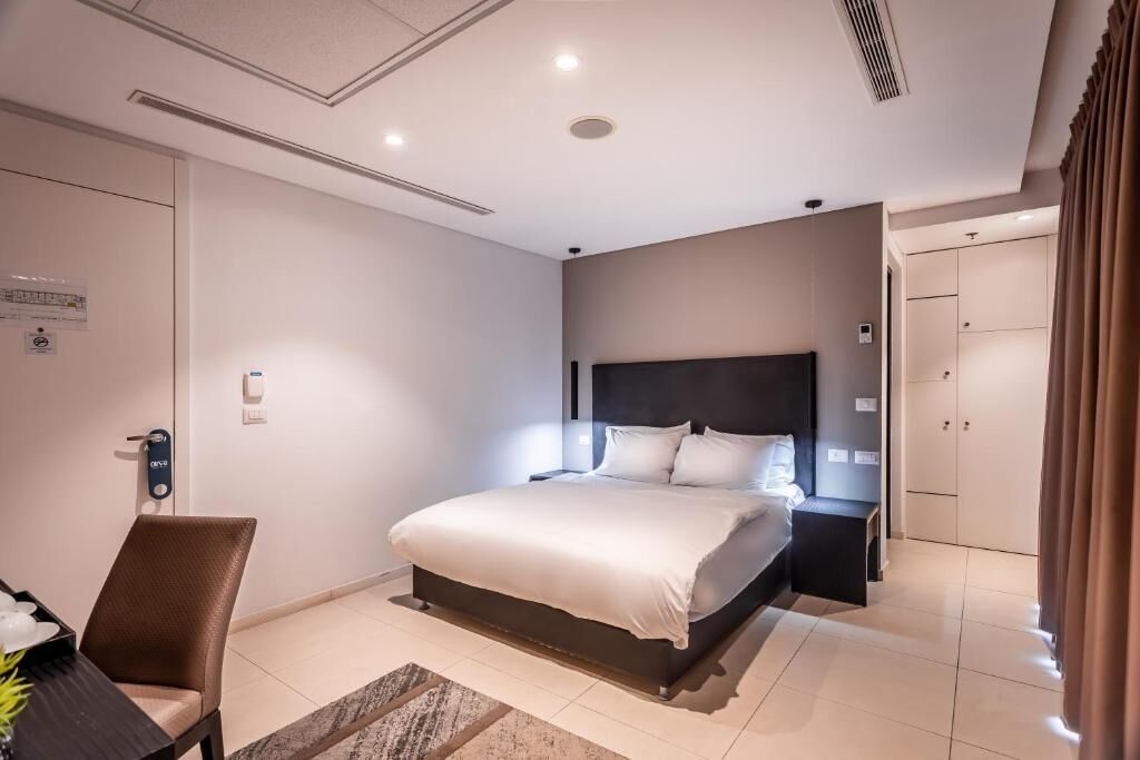 A modern hotel room with a large bed, a desk with a chair, a wardrobe, and a bathroom door.