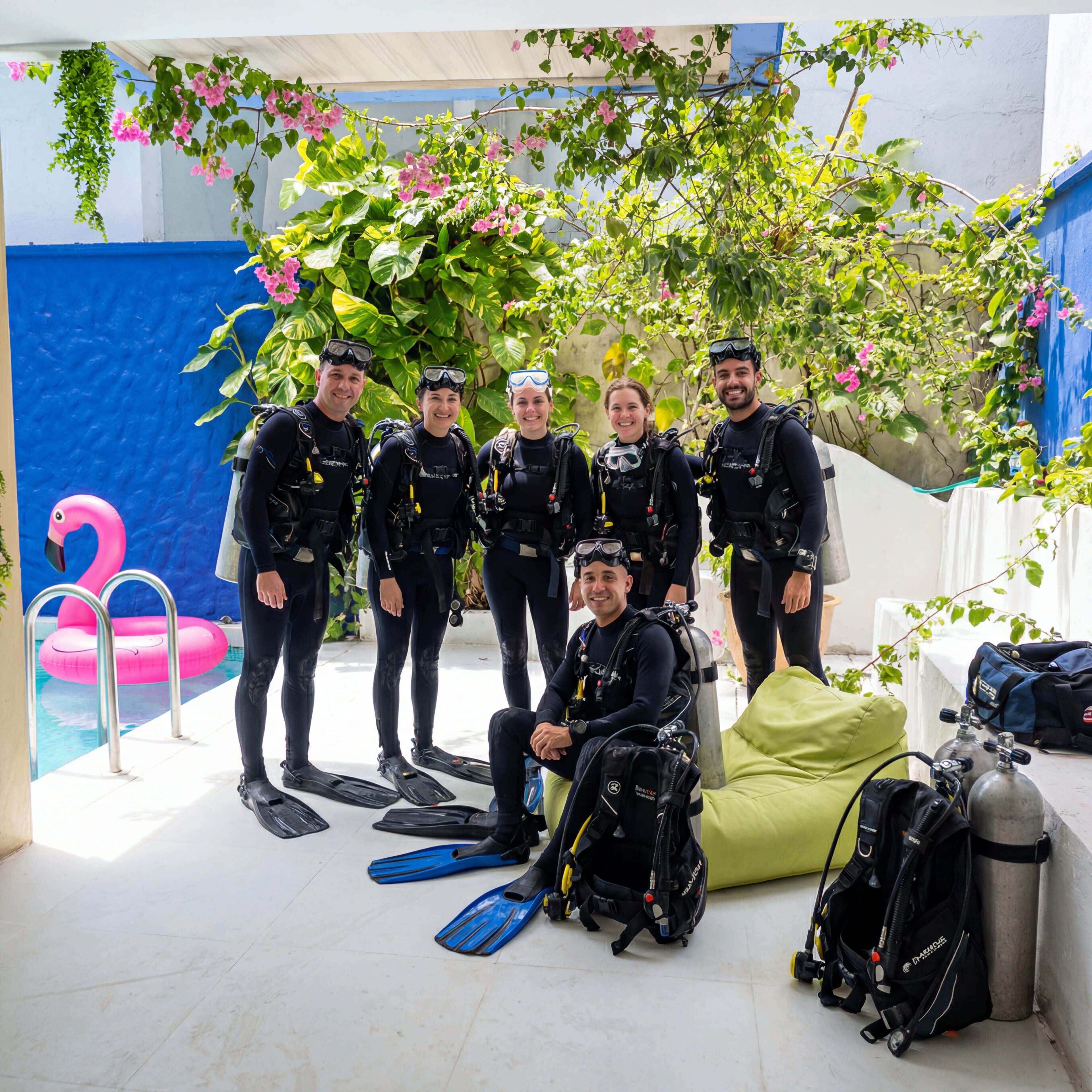 A group of divers in a covered outdoor area with a pool, diving equipment, and a pink flamingo float.