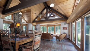 A rustic interior with wooden beams, a stone fireplace, wicker furniture, and large windows offering views of the outdoors.
