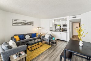 A modern living room with a grey sofa, a coffee table, and a dining area with a black table. - Includes Xplorie Activities and Linens. PYRAMID - 1BR oceanfront condo w/outdoor pool. (Ocean City)