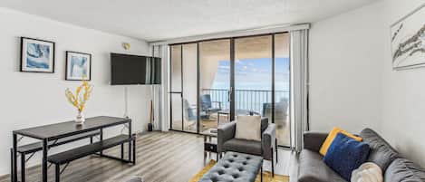 Un salon moderne avec un canapé, une table basse et un téléviseur. Il y a un balcon avec vue sur la mer.
