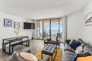 A modern living room with a sofa, a coffee table, and a TV. There is a balcony with a view of the sea. - Includes Xplorie Activities and Linens. PYRAMID - 1BR oceanfront condo w/outdoor pool. (Ocean City)