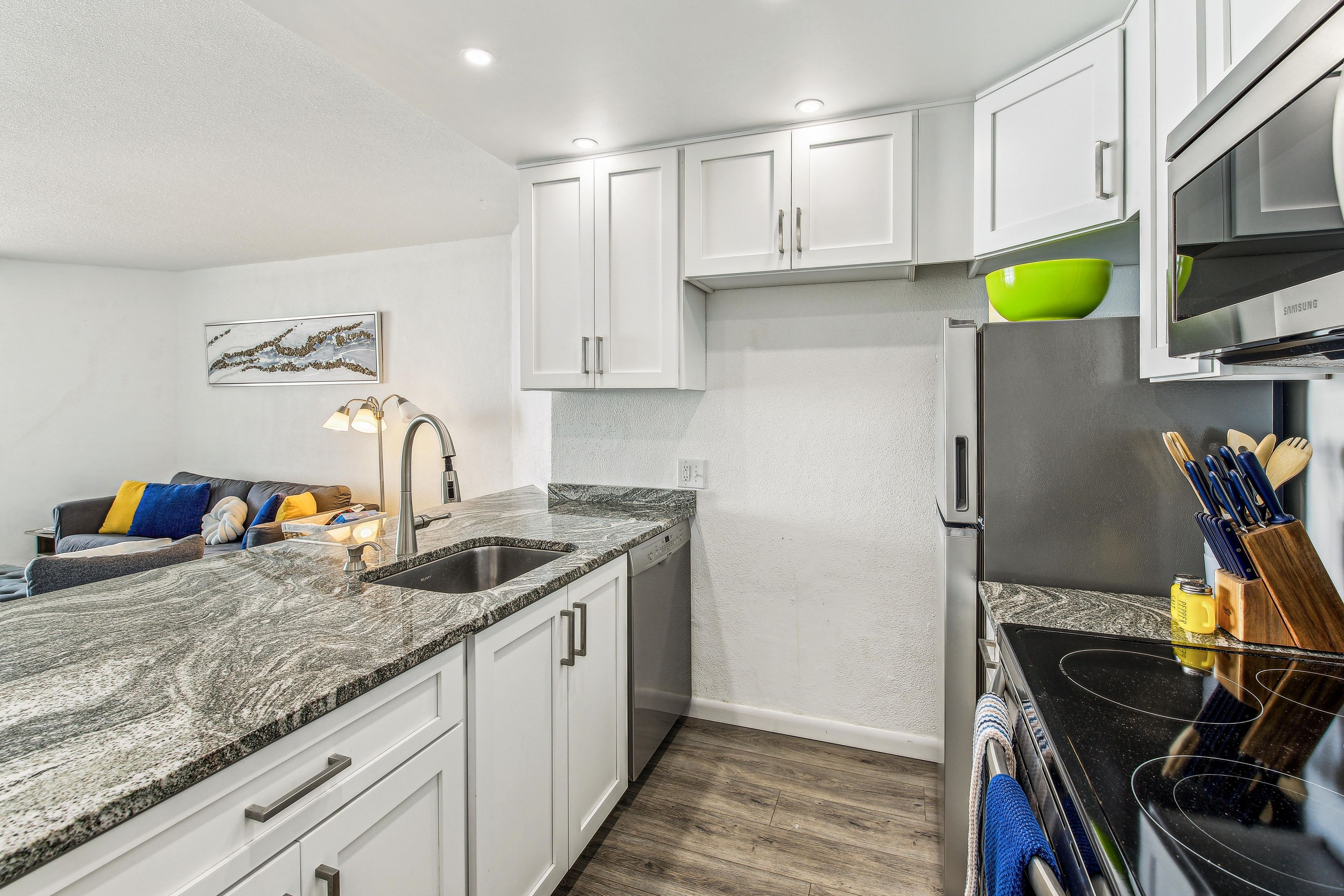 A modern kitchen with white cabinets, a granite countertop, a stainless steel refrigerator, and a dining area with a couch and pillows.