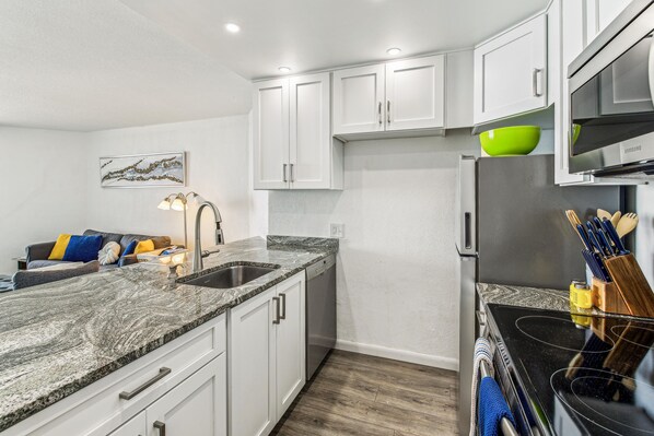 A modern kitchen with white cabinets, a granite countertop, a stainless steel refrigerator, and a dining area with a couch and pillows. - Includes Xplorie Activities and Linens. PYRAMID - 1BR oceanfront condo w/outdoor pool. (Ocean City)