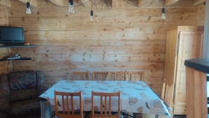 A wooden cabin interior with a dining table, chairs, a sofa, and a television on a shelf. - Chalet "Les Mouflons" - Le Clos - Sleeps 8, 5 minutes walk from the ski slopes (Crest-Voland)