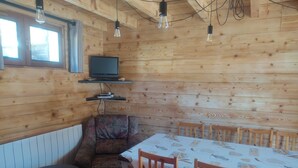 A wooden cabin interior with a dining table, chairs, a sofa, a television on a shelf, and hanging light bulbs. - Chalet "Les Mouflons" - Le Clos - Sleeps 8, 5 minutes walk from the ski slopes (Crest-Voland)
