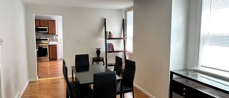 A spacious living room with hardwood flooring, a dining table with chairs, a bookshelf, and a view of the kitchen through an open doorway.