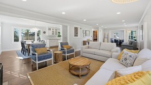 Amplia sala de estar con un sofá grande, una mesa de centro y una mesa de comedor con sillas. Se observa una cocina al fondo con armarios blancos y un rincón de comedor con una silla a rayas.