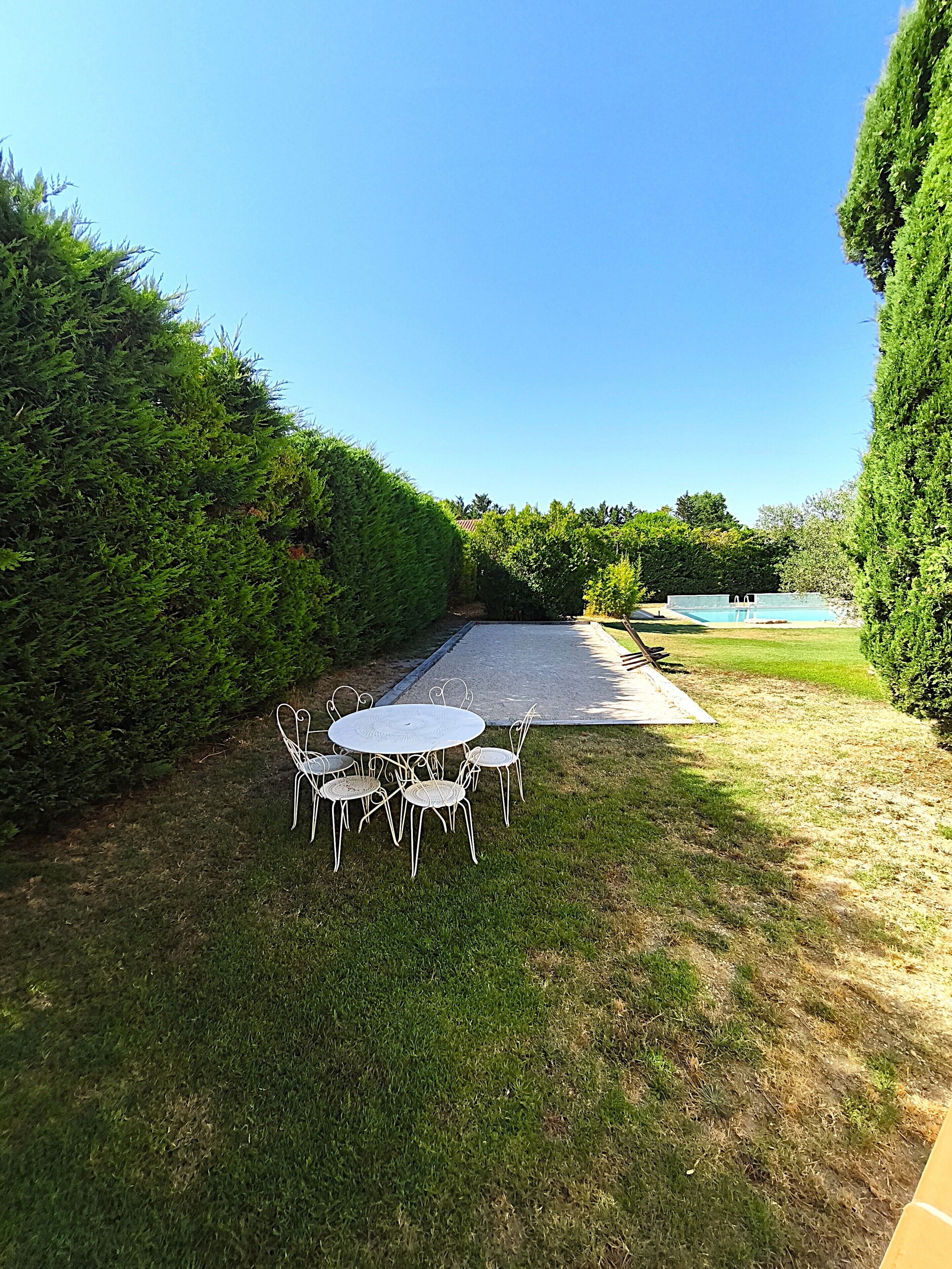 A patio with a round table and chairs, a swimming pool, and a hedge-lined pathway.