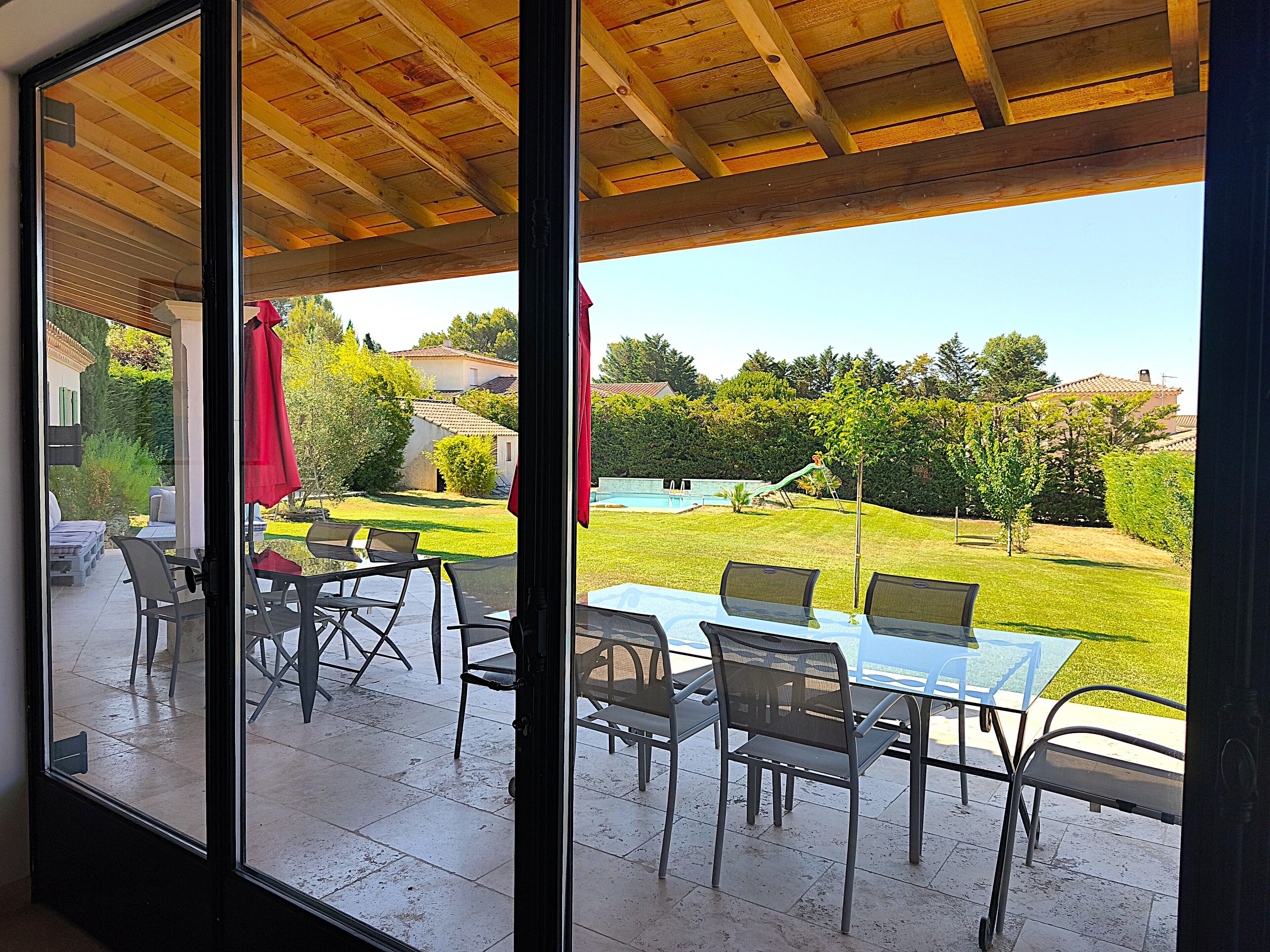 A covered patio with a glass table and chairs, a red umbrella, and a view of a backyard with a pool and trees.