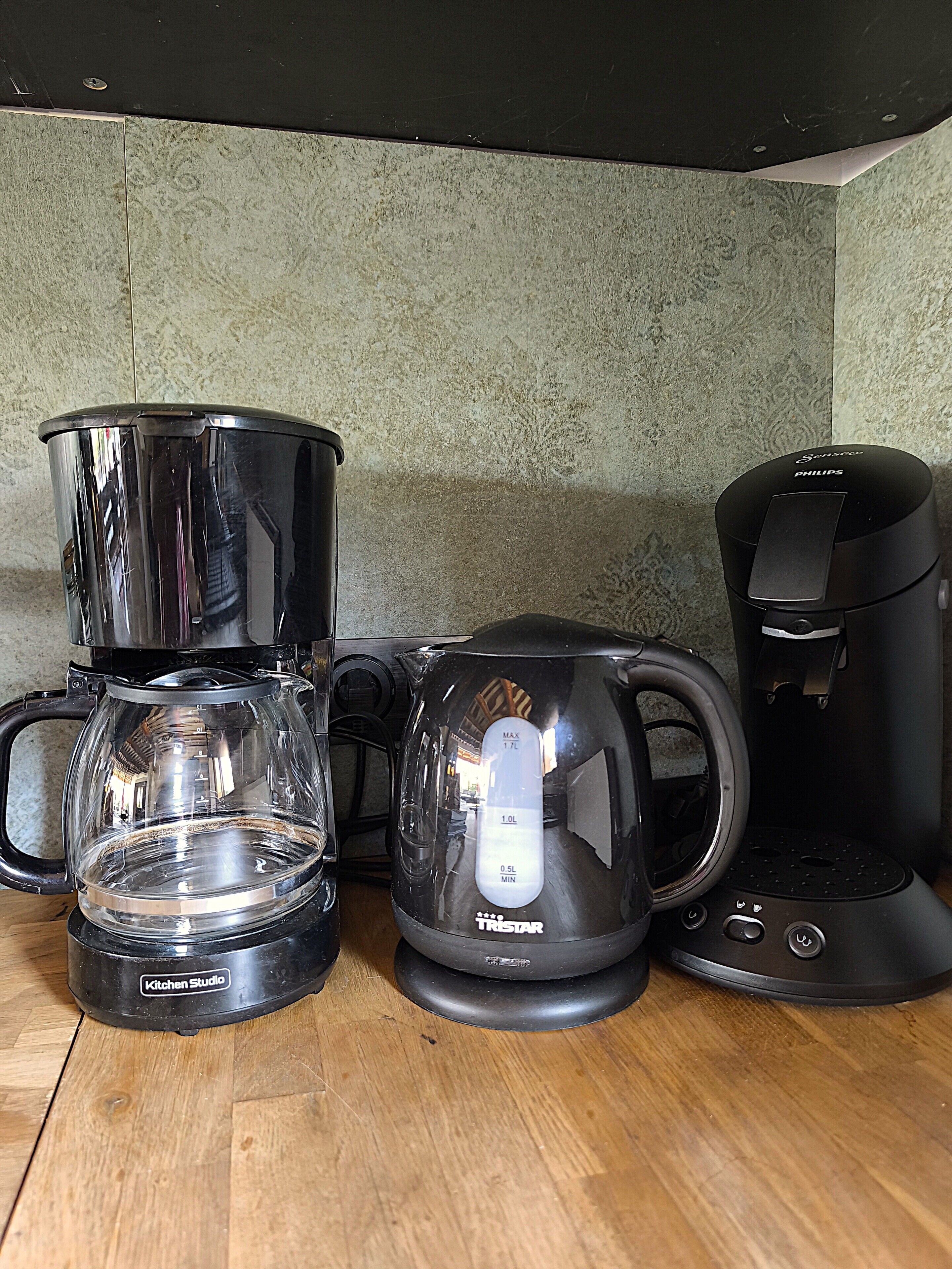 A coffee maker, a glass carafe, and a coffee pot on a wooden table against a textured wall.