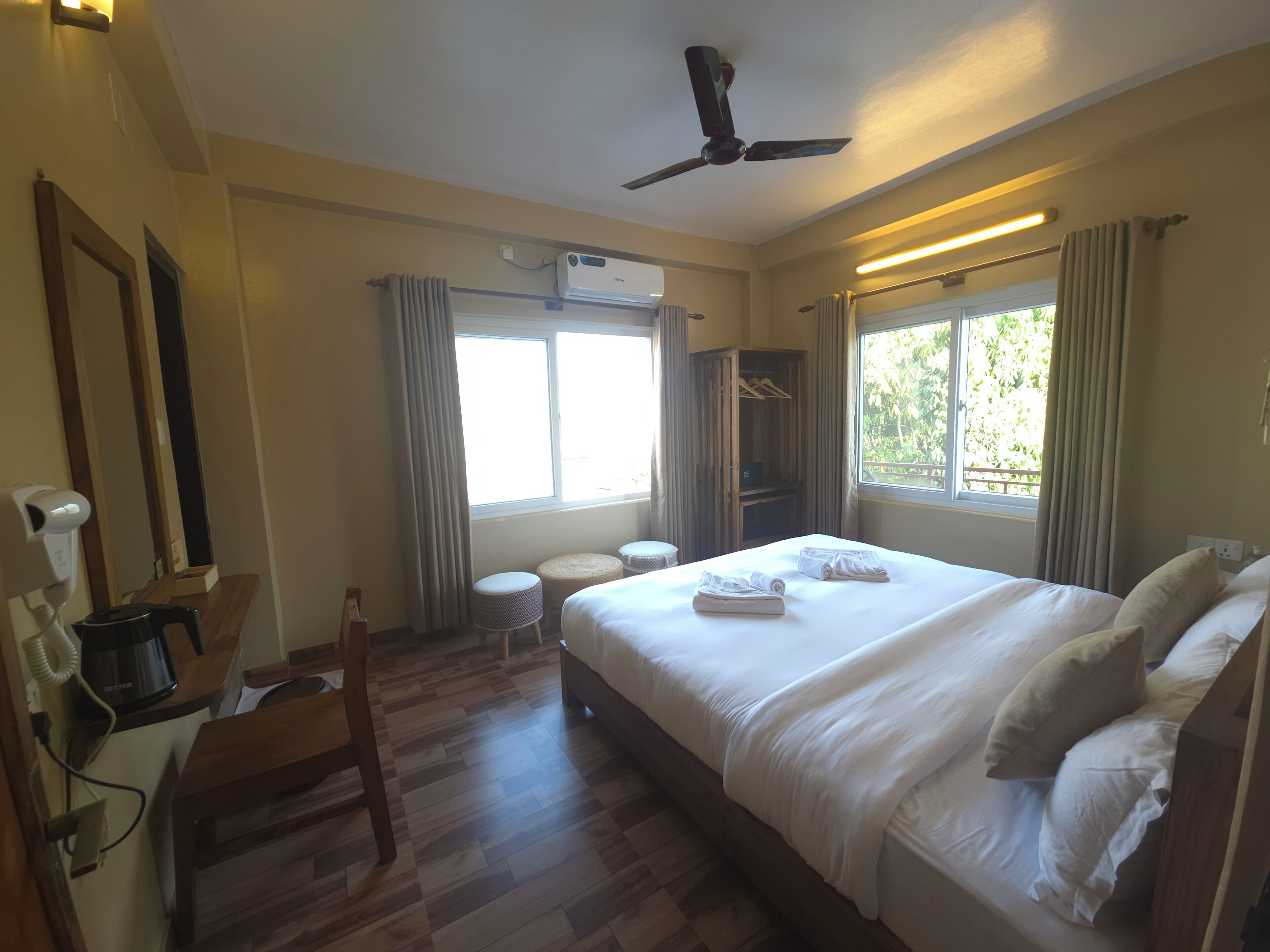 A hotel room with a bed, a desk with a coffee maker, a wardrobe, a ceiling fan, and a window with curtains.