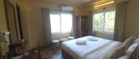 A hotel room with a bed, a desk with a coffee maker, a wardrobe, a ceiling fan, and a window with curtains.