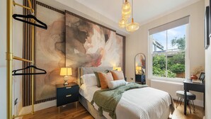 Une chambre avec un grand tableau abstrait, un lit recouvert de plusieurs coussins, un bureau avec une chaise et une fenêtre donnant sur un paysage verdoyant.