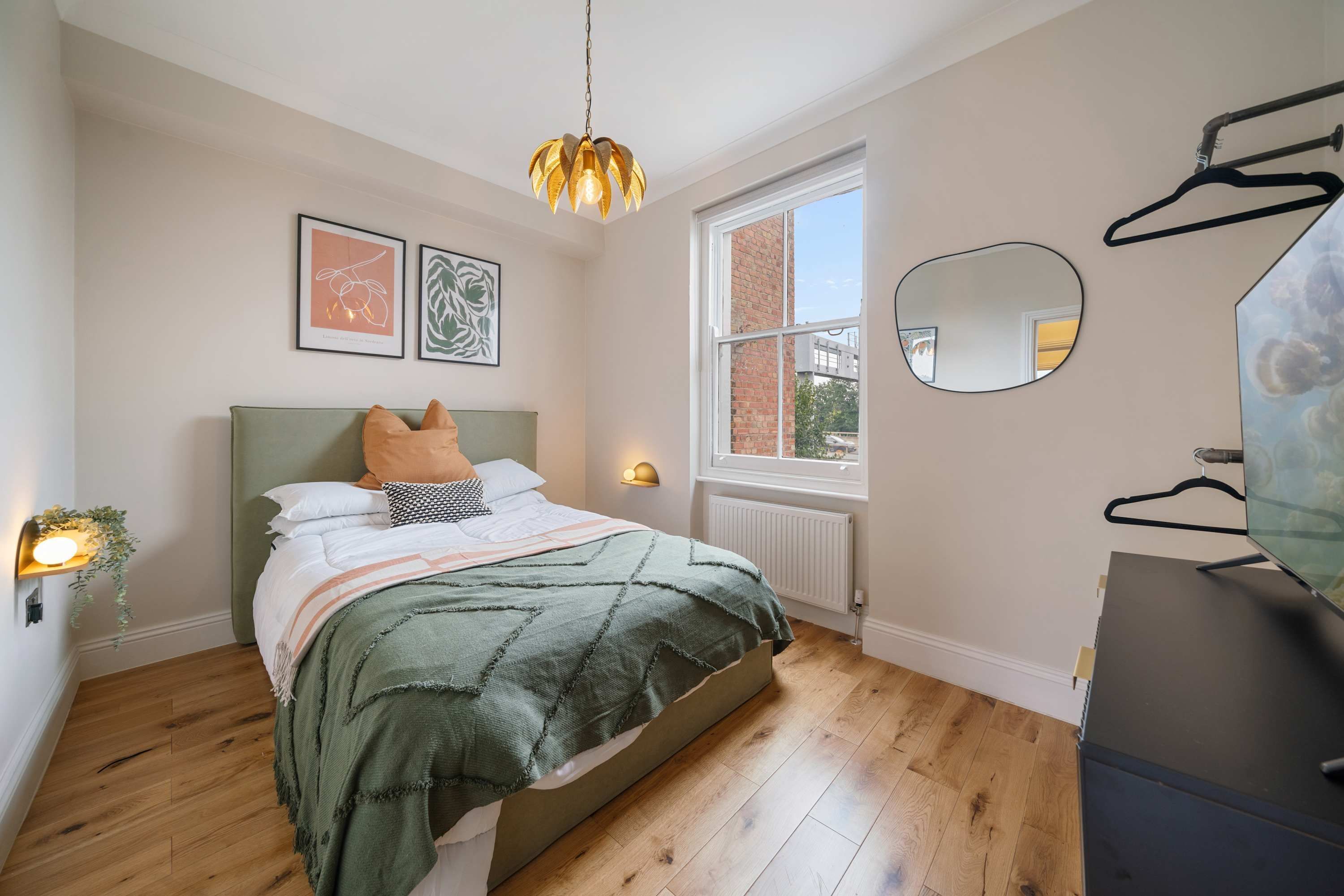 Ein Schlafzimmer mit Bett, Pendelleuchte, Fenster, Heizkörper und Spiegel an der Wand.