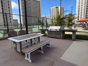 Un patio al aire libre con mesa y sillas, una parrilla y una cancha de tenis al fondo.