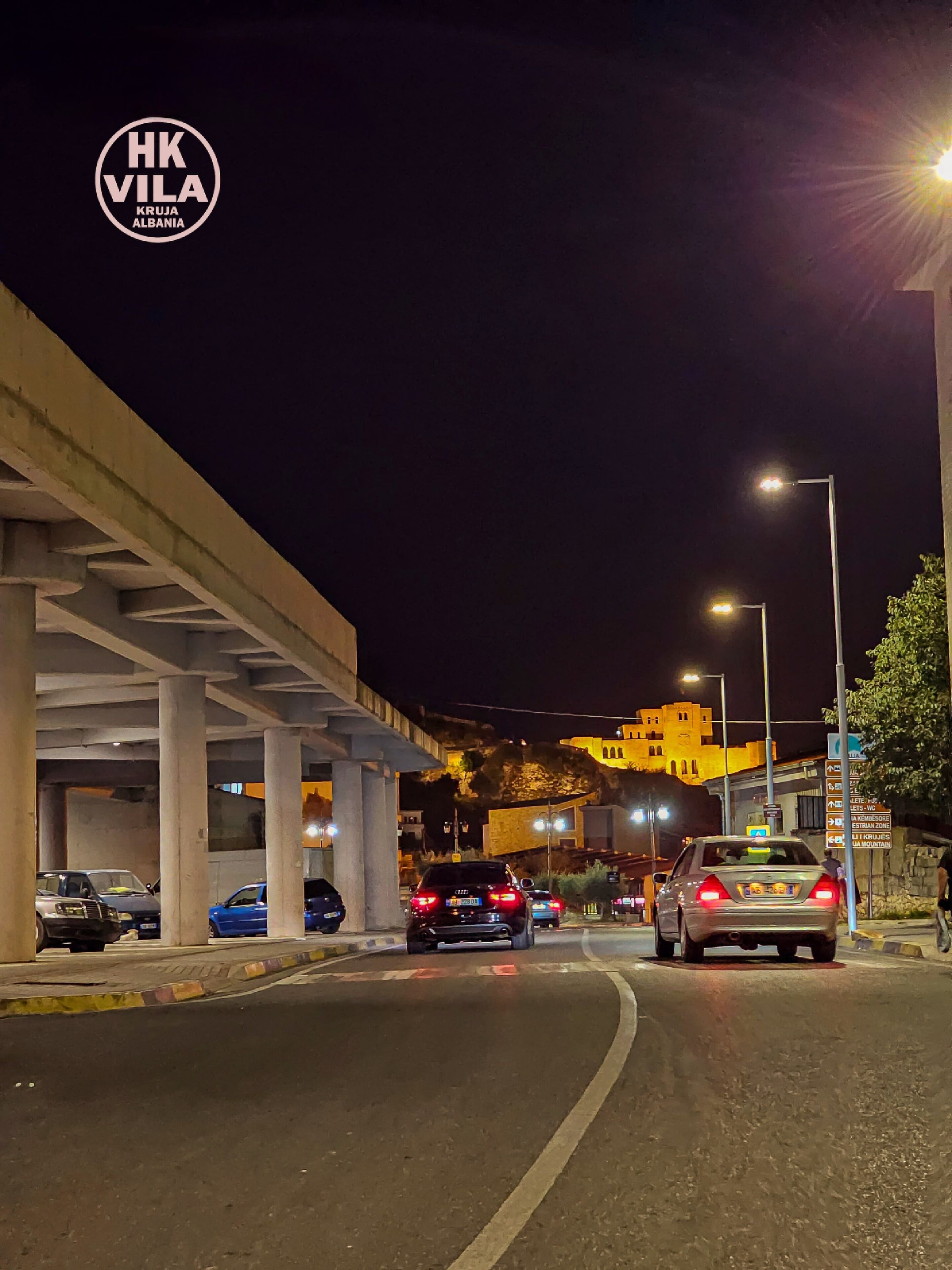 A nighttime street scene with cars, a bridge, and illuminated buildings.