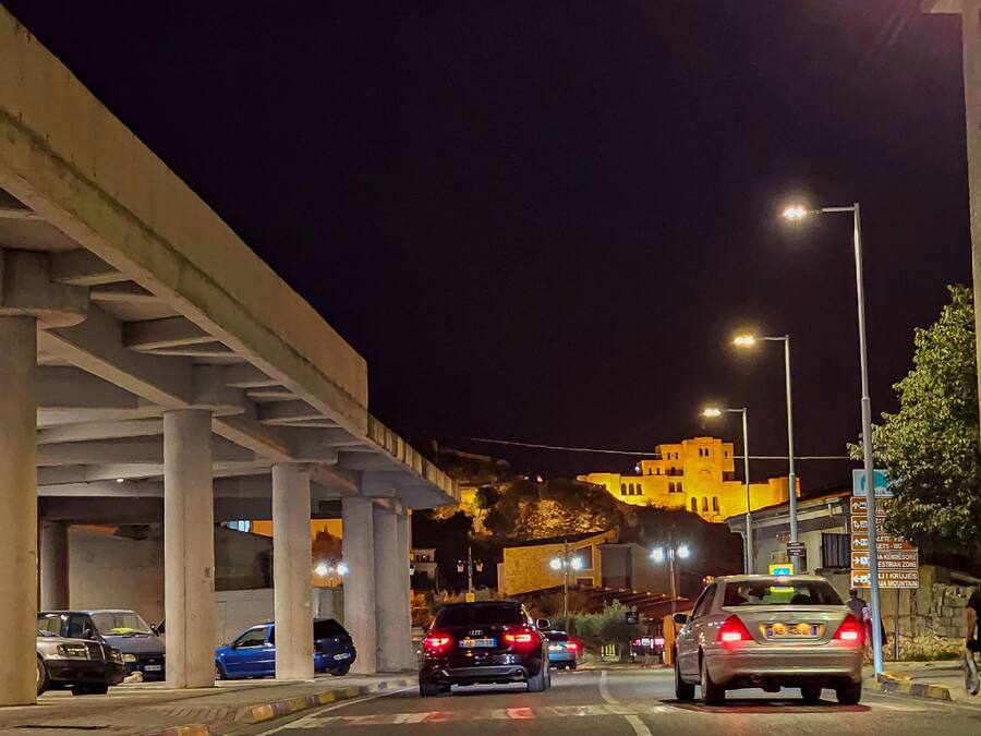 A nighttime street scene with cars, a bridge, and illuminated buildings.