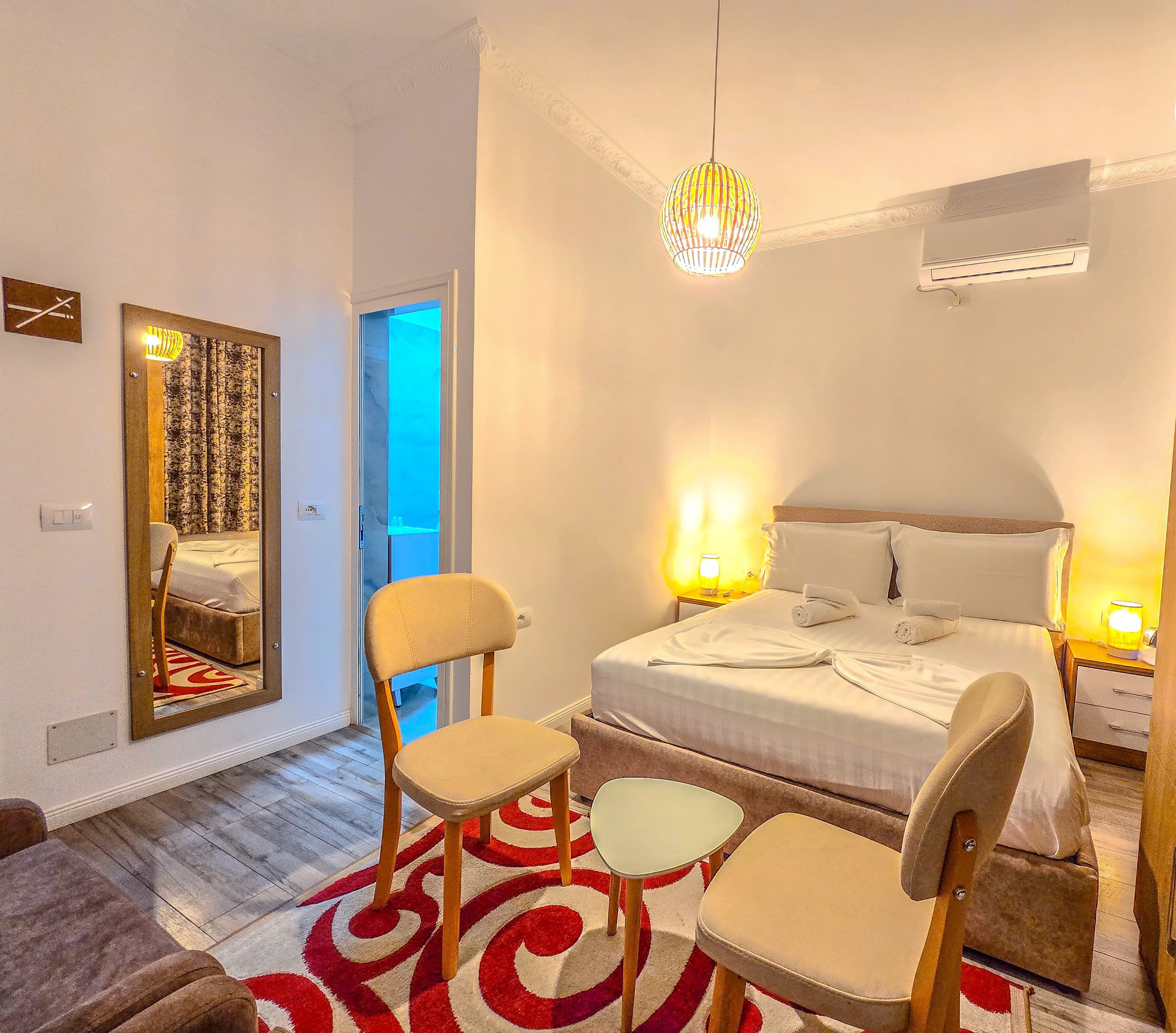 A hotel room with a bed, two chairs, a mirror, a chandelier, and a red and white patterned rug.