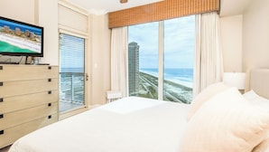 A hotel room with a large bed, a ceiling fan, a TV on a dresser, and a view of the beach and ocean.